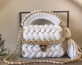 May include: White crocheted handbag with a gold chain strap and a gold clasp. The bag has a woven texture and a tassel.