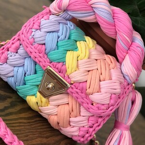 May include: A rainbow-colored crocheted handbag with a braided handle and a gold clasp. The bag has a long, braided strap.