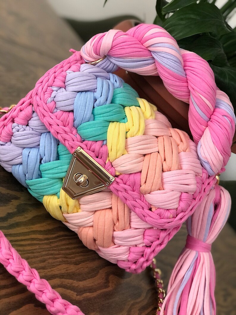 May include: A rainbow-colored crocheted handbag with a braided handle and a gold clasp. The bag has a long, braided strap.