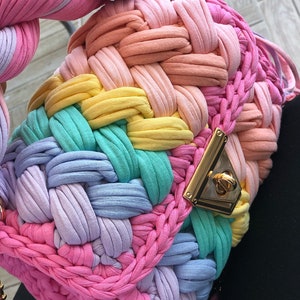 May include: A rainbow-colored crocheted bag with a gold clasp. The bag is made of thick, colorful yarn and has a woven texture.
