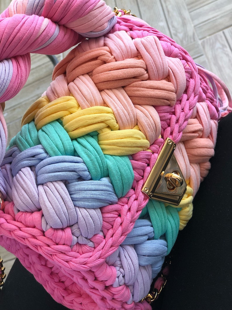 May include: A rainbow-colored crocheted bag with a gold clasp. The bag is made of thick, colorful yarn and has a woven texture.