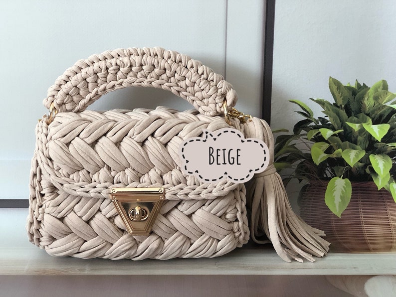 Peut inclure: Sac &agrave; main en crochet beige avec une fermeture dor&eacute;e et un pompon. Le sac a un design tiss&eacute; et une poign&eacute;e sup&eacute;rieure.
