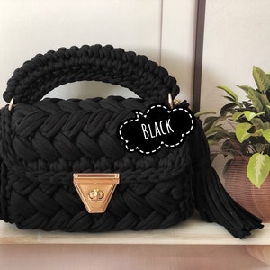 Peut inclure: Sac &agrave; main en crochet noir avec un fermoir dor&eacute; et un pompon noir. Le sac a un design tress&eacute; et une poign&eacute;e sup&eacute;rieure.