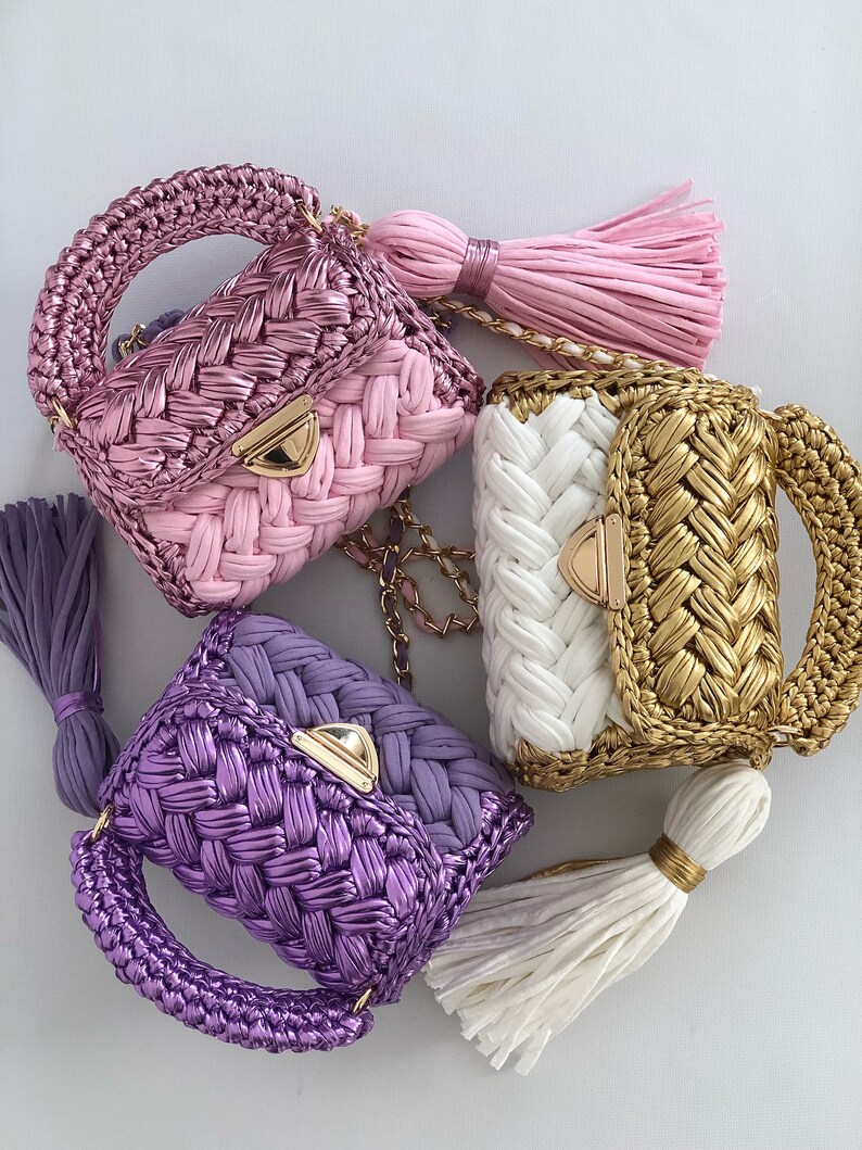 May include: Three small crocheted handbags with gold chain straps and tassels. The handbags are pink, white and gold, and purple.
