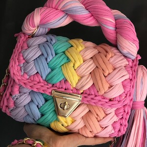 May include: A rainbow-colored crocheted handbag with a braided handle and a gold clasp. The bag has a long, tassel-like strap.