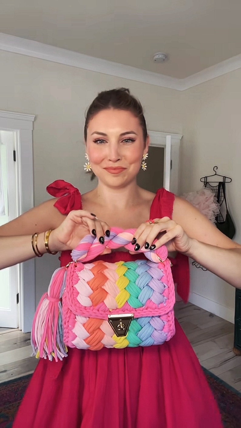 May include: A woman wearing a pink dress and holding a rainbow crocheted handbag with a gold clasp and tassel.