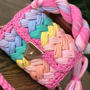 May include: A colorful crocheted handbag with a braided handle and a gold clasp. The bag is made of multiple colors of yarn, including pink, purple, blue, green, and yellow.