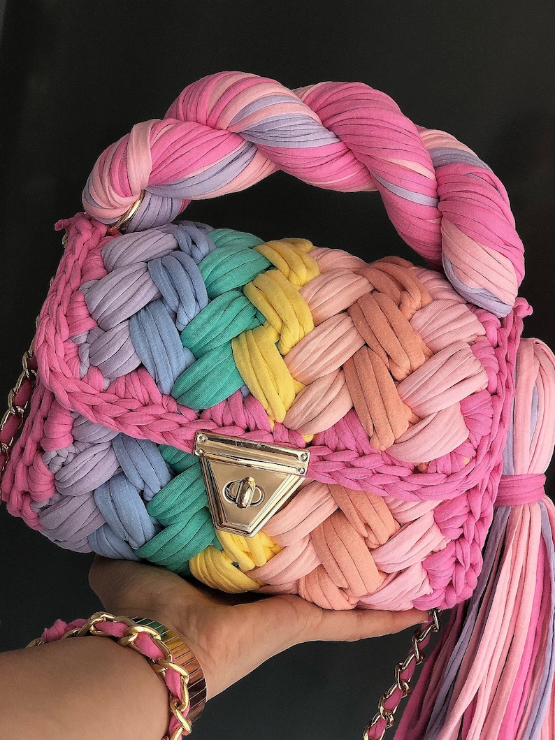 May include: A rainbow-colored crocheted handbag with a braided handle and a gold clasp. The bag has a long, colorful tassel.