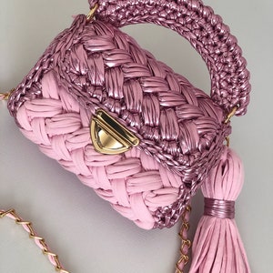 May include: A pink crocheted handbag with a gold chain strap and a tassel. The bag has a woven design and a metallic finish.