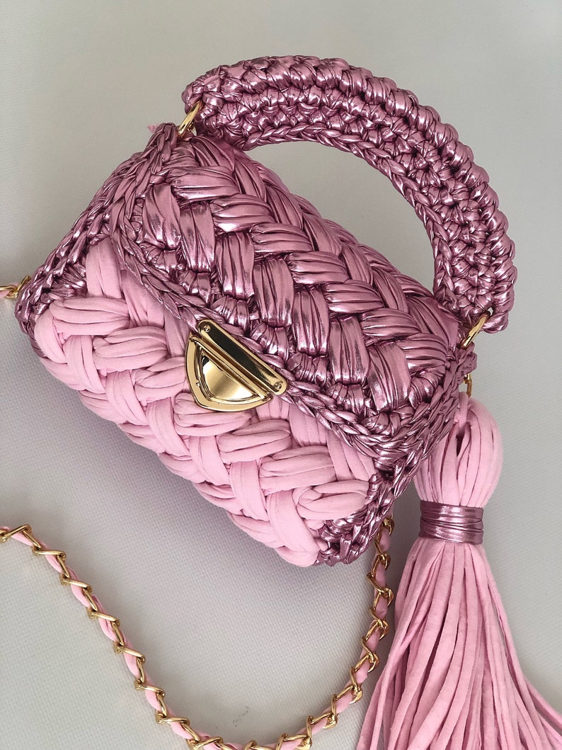 May include: A pink crocheted handbag with a gold chain strap and a tassel. The bag has a woven design and a metallic finish.