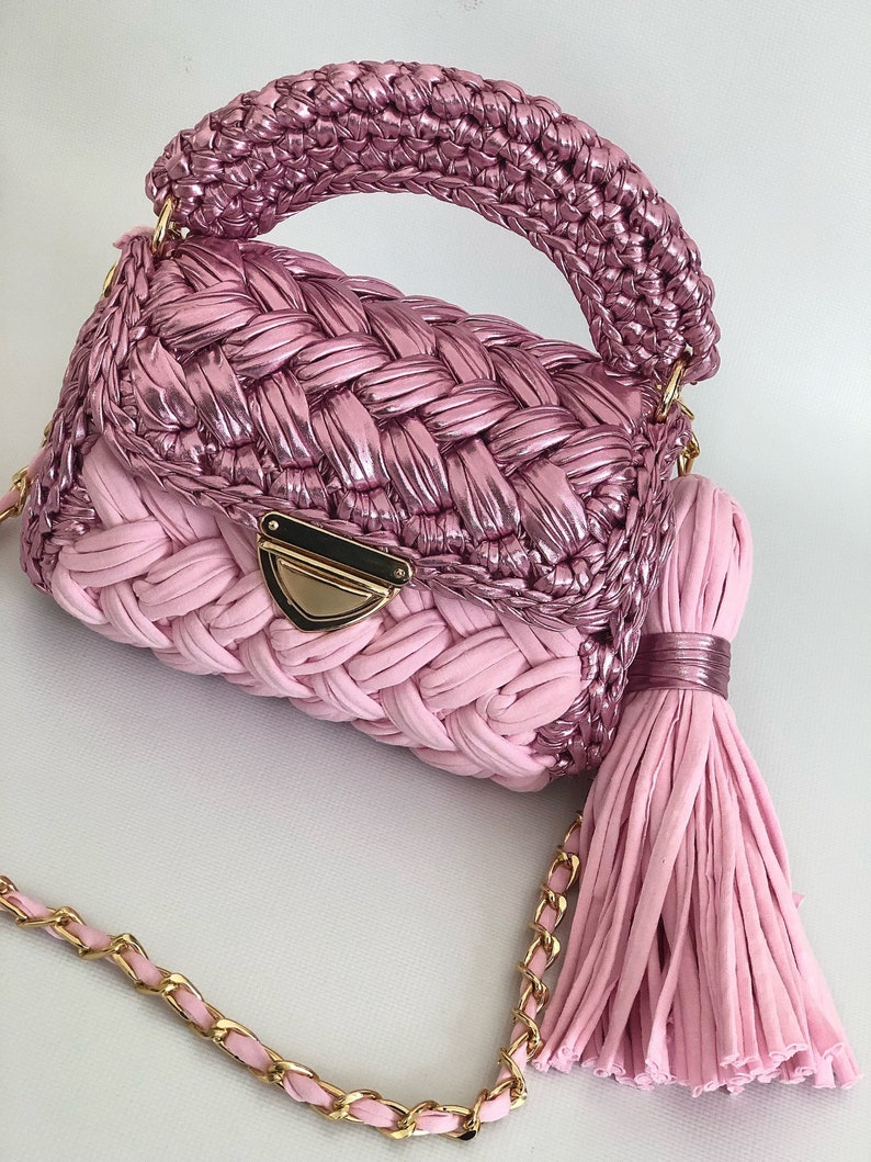 May include: A pink crocheted handbag with a gold chain strap and a tassel. The bag has a woven design and a top handle.