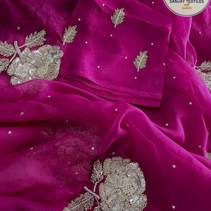 May include: A vibrant fuchsia saree with intricate silver floral embroidery and delicate leaf accents. The fabric is draped, showcasing the detailed embellishments and scattered sequins. The Sanjay Textiles logo is visible in the upper right corner.