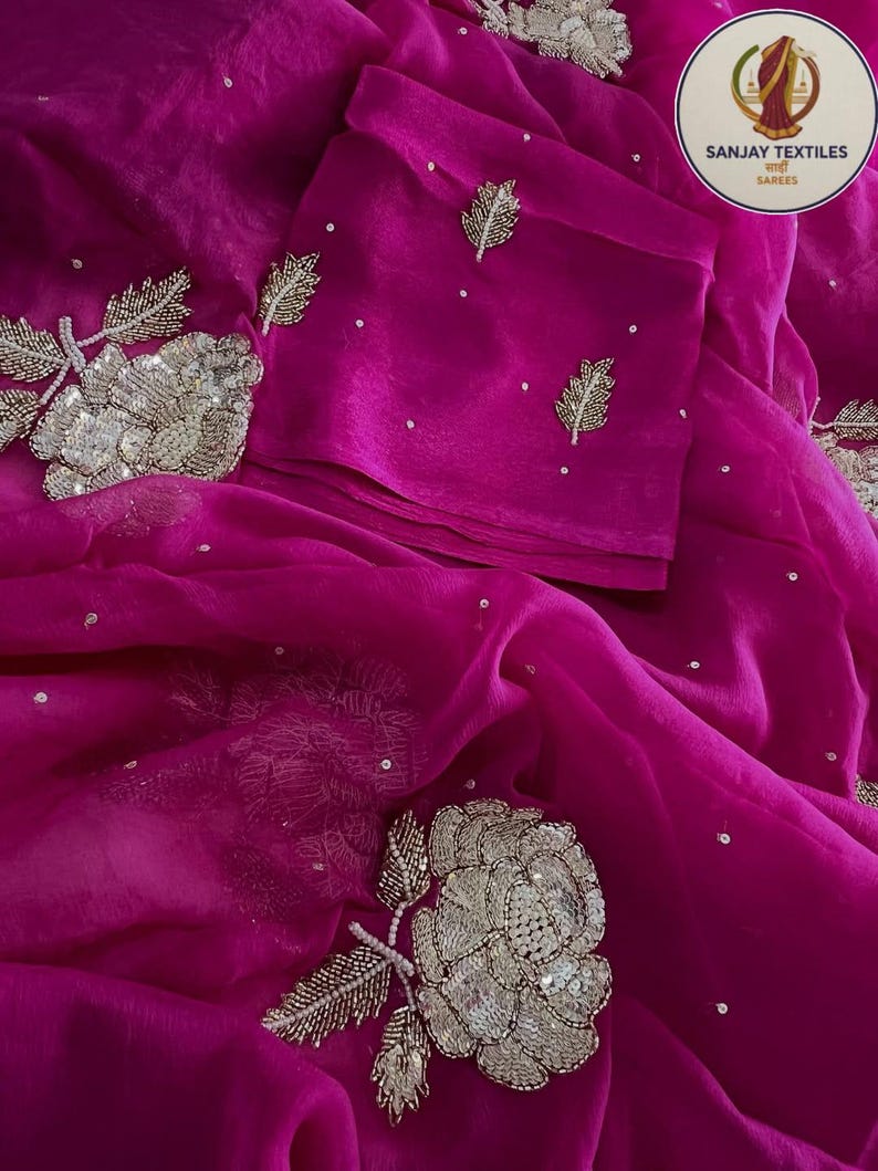 May include: A vibrant fuchsia saree with intricate silver floral embroidery and delicate leaf accents. The fabric is draped, showcasing the detailed embellishments and scattered sequins. The Sanjay Textiles logo is visible in the upper right corner.