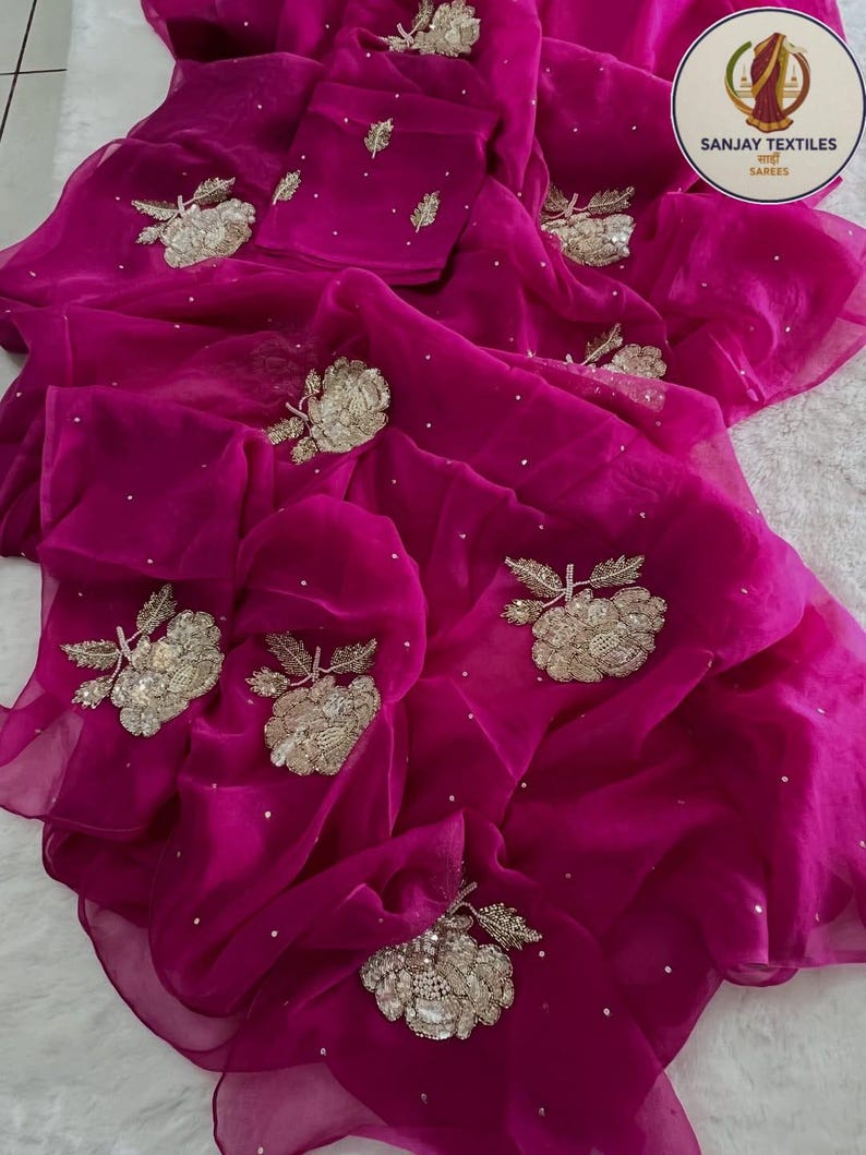 May include: A vibrant fuchsia saree with intricate gold floral embroidery and scattered white sequins. The sheer fabric drapes elegantly, showcasing the detailed craftsmanship. The saree features a matching blouse piece, perfect for special occasions.