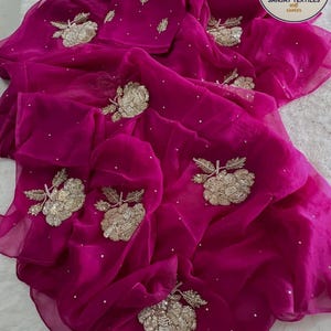 May include: A vibrant fuchsia saree with intricate gold floral embroidery and scattered white sequins. The sheer fabric drapes elegantly, showcasing the detailed craftsmanship. The saree features a matching blouse piece, perfect for special occasions.