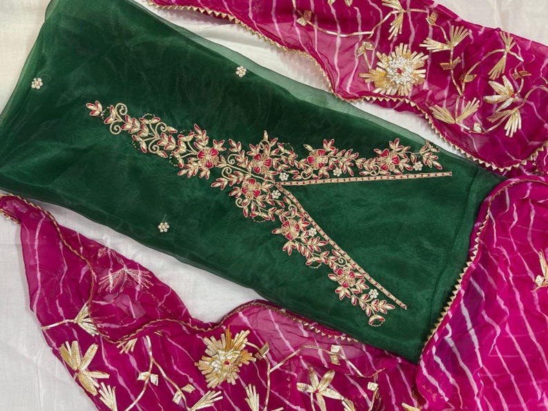 May include: A dark green fabric with intricate floral embroidery in gold and red. The design is complemented by a vibrant pink fabric with gold accents. The ensemble suggests a traditional garment, possibly a dress or a set.