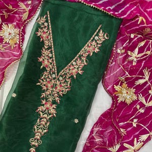 May include: A dark green salwar kameez with intricate floral embroidery in red and gold thread. The neckline and bodice feature detailed patterns. A pink dupatta with gold accents and a striped pattern complements the outfit. The fabric appears sheer and lightweight.