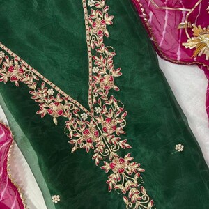 May include: Dark green fabric with intricate floral embroidery in red and gold. The embroidery forms a V-neck design and floral patterns. A pink and gold patterned fabric is visible in the background.