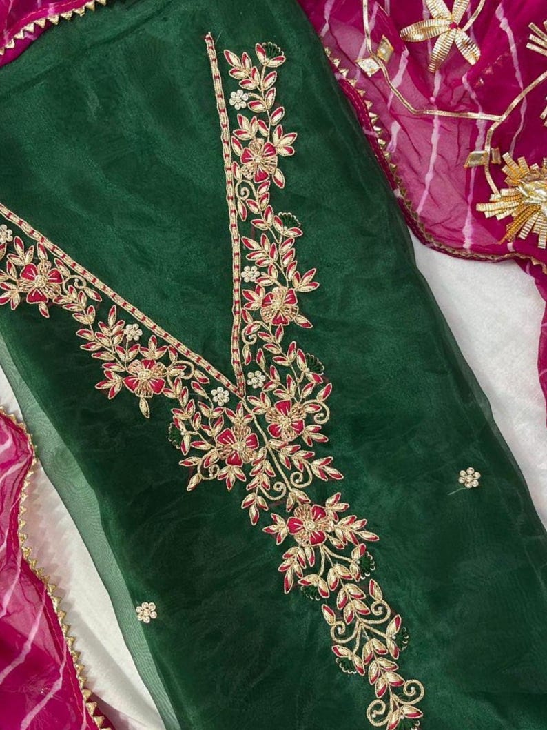 May include: Dark green fabric with intricate floral embroidery in red and gold. The embroidery forms a V-neck design and floral patterns. A pink and gold patterned fabric is visible in the background.