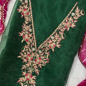 May include: Emerald green fabric with intricate floral embroidery in red, gold, and white. The embroidery forms a V-neck design and is accented with delicate leaf patterns. A pink and white patterned scarf is visible in the background.