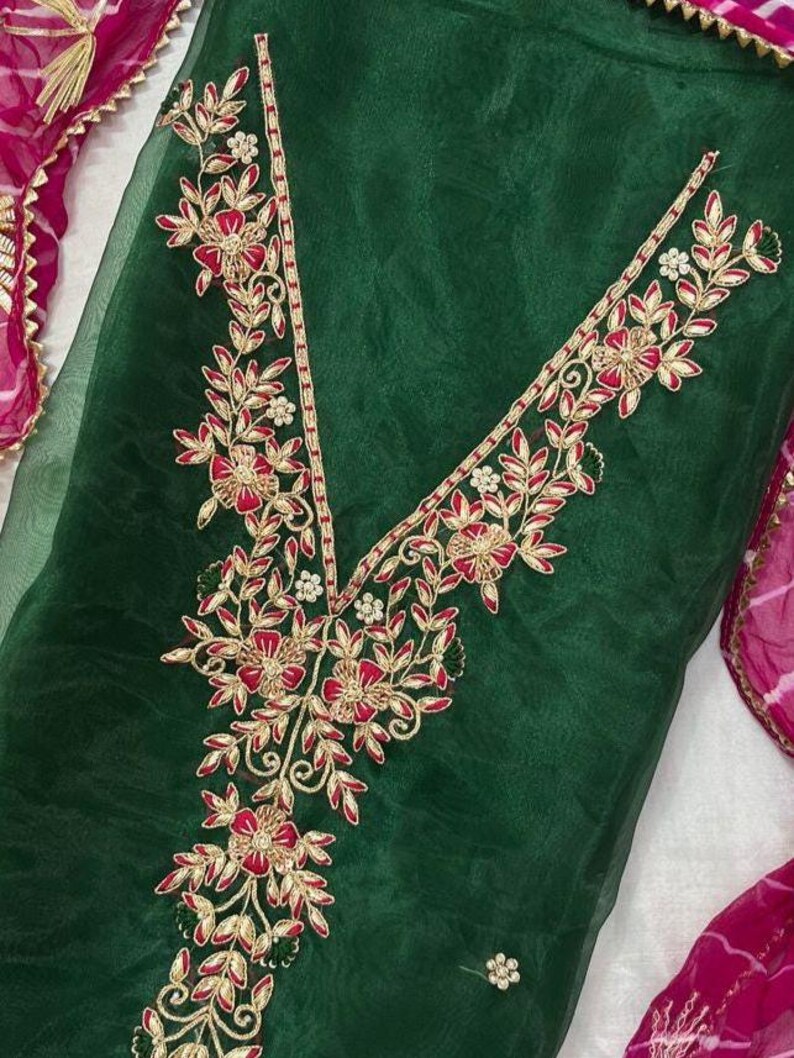 May include: Emerald green fabric with intricate floral embroidery in red, gold, and white. The embroidery forms a V-neck design and is accented with delicate leaf patterns. A pink and white patterned scarf is visible in the background.