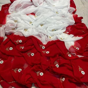 May include: A red and white fabric with floral embroidery. The fabric is draped and folded, with the red sections featuring small, embroidered flowers with green centers and gold accents. The white sections also have similar embroidery.