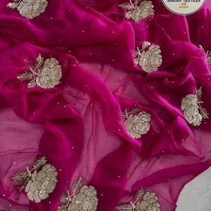 May include: A vibrant fuchsia fabric with intricate floral embroidery. The embroidered flowers are detailed with silver thread and sequins. The fabric is draped, creating soft folds and a luxurious appearance. The image also includes the text "SANJAY TEXTILES" and "SAREES".