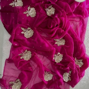 May include: A vibrant magenta saree with intricate floral embroidery. The sheer fabric drapes elegantly, adorned with detailed gold and silver embellishments. The saree features a flowing design, perfect for special occasions.