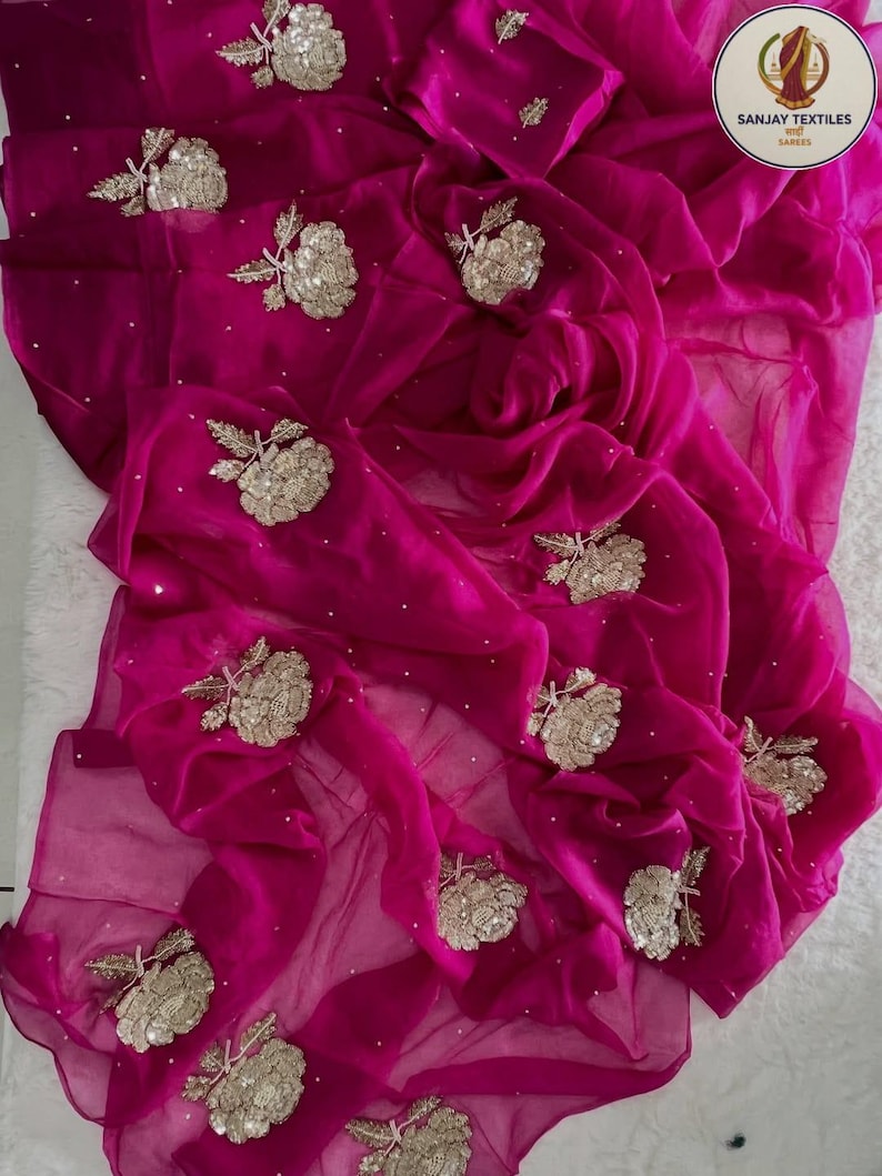 May include: A vibrant magenta saree with intricate floral embroidery. The sheer fabric drapes elegantly, adorned with detailed gold and silver embellishments. The saree features a flowing design, perfect for special occasions.