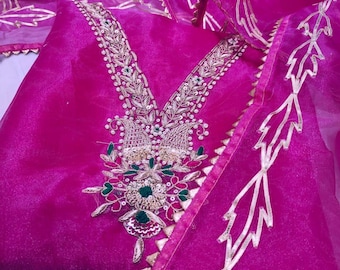 May include: A vibrant fuchsia-colored garment with intricate gold embroidery and embellishments. The neckline and edges are adorned with detailed patterns, and the sheer fabric adds a delicate touch. The design features floral and leaf motifs.