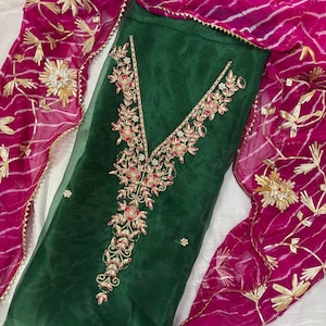 May include: A dark green, sheer fabric garment with intricate floral embroidery in red, gold, and white. A bright pink dupatta with gold accents and a striped pattern is draped around the garment. The ensemble is likely a traditional outfit.