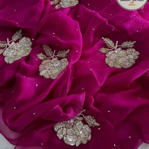 May include: A vibrant magenta fabric with intricate silver floral embroidery and scattered silver sequins. The fabric is draped, showcasing its flowing texture and the detailed craftsmanship of the embellishments. The Sanjay Textiles logo is in the upper right corner.