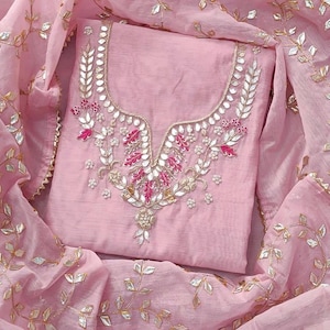 May include: A light pink embroidered salwar kameez set. The top features intricate floral embroidery in silver, white, and pink around the neckline. The matching dupatta is a sheer fabric with silver leaf accents.