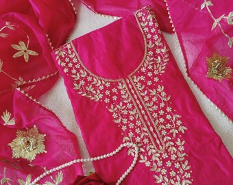 May include: A vibrant fuchsia-colored salwar kameez set. The tunic features intricate gold embroidery around the neckline and down the center. A matching dupatta and a pearl necklace with a red rose complete the ensemble.