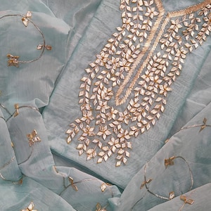 May include: A pale blue two-piece outfit featuring gold embroidery. The neckline and sleeves are decorated with a leaf design. The fabric has a delicate, flowing texture, and the design includes small, gold embellishments.