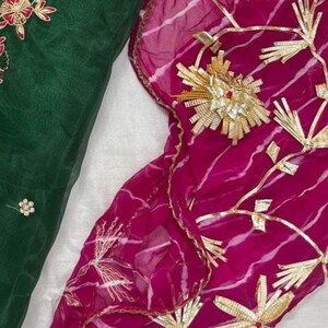 May include: Close-up of two pieces of fabric. One is a deep emerald green with floral embroidery in red and gold. The other is a vibrant magenta with gold floral and geometric embellishments. The fabrics appear to be lightweight and flowing.
