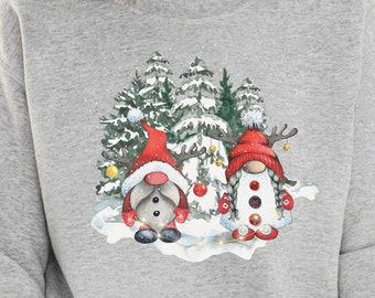 Op de afbeelding: Een lichtgrijs sweatshirt met een kerstontwerp. De afbeelding toont twee kabouters met rode mutsen en wanten, in een besneeuwde scène met dennenbomen. De kabouters hebben knoopogen en zijn versierd met kerstballen.