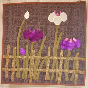 Könnte beinhalten: Ein gesteppter Wandbehang mit braunem Hintergrund, der eine stilisierte Darstellung eines Gartens mit lila und weißen Blumen hinter einem braunen Zaun zeigt.