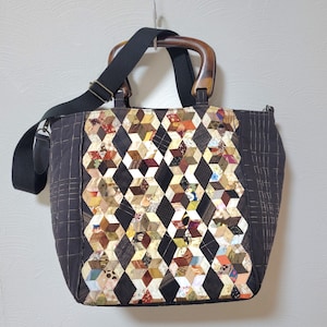 Puede incluir: Bolso tote hecho a mano con un diseño de patchwork geométrico en tonos marrón, beige y negro. El bolso tiene asas de madera y una correa para el hombro negra. Los laterales del bolso son de color marrón oscuro liso.