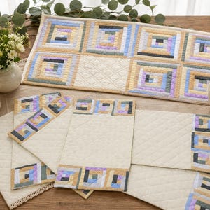 Op de afbeelding: Een rechthoekige gewatteerde tafelloper en bijpassende placemats met een log cabin blokpatroon in tinten beige, lichtblauw en lavendel. De stoffen delen zijn gerangschikt in concentrische vierkanten met crèmekleurige gewatteerde delen.