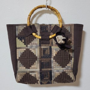 Puede incluir: Una bolsa de mano de lona marrón con un asa de bambú. La bolsa está hecha de cuadrados de tela en diferentes tonos de marrón y beige. Una pequeña flor de tela marrón está unida al asa.