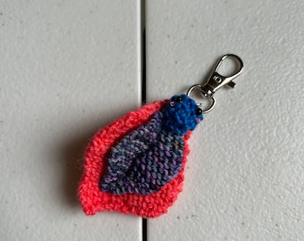 Könnte beinhalten: Ein kleiner, gestrickter Schlüsselanhänger in Form eines Blattes. Das Blatt besteht aus zwei Farben: einem dunkelblauen und violetten Garn und einem leuchtend rosa Garn. Der Schlüsselanhänger hat eine silberne Metallschaufel.