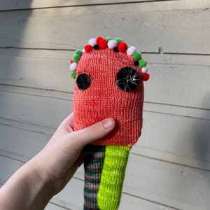 May include: A hand-knitted plush toy with a red body, green and white pom-poms, and black button eyes.