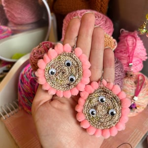 Puede incluir: Un par de pendientes de crochet rosa y marrón con tres ojos blancos en cada pendiente.