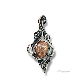 May include: A handcrafted pendant featuring a teardrop-shaped, speckled, brown gemstone set within a silver-toned, ornate wire design. The pendant has a unique, artistic style, perfect for adding a touch of elegance to any outfit.