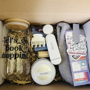 Puede incluir: Una caja de cartón marrón llena de una variedad de artículos, que incluyen un frasco de vidrio con una tapa de madera que dice "Let's go book shopping", un peine blanco, una crema de manos blanca, una unidad USB blanca, una toalla gris y un marcapáginas con el texto "Care for Quitter's Characters".