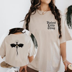 May include: Beige t-shirt with the text "Violent Little Thing" in black gothic font on the front. The back of the shirt features a black dragon graphic with a sunburst halo. The shirt is a casual, oversized fit.