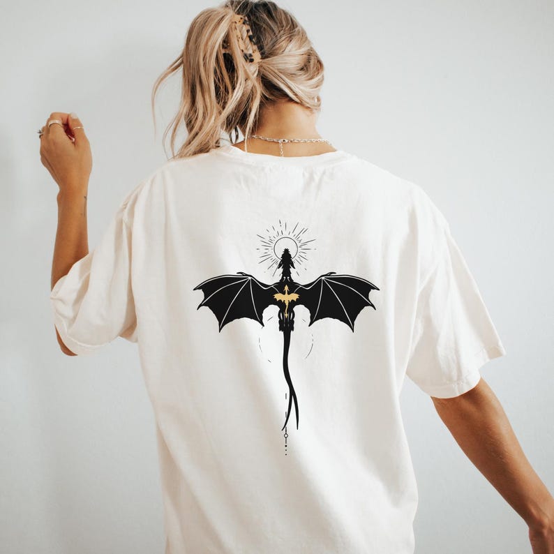 May include: Cream-colored t-shirt featuring a black dragon graphic on the back. The dragon has outstretched wings and a gold-colored chest. The design includes a sunburst and a long tail.
