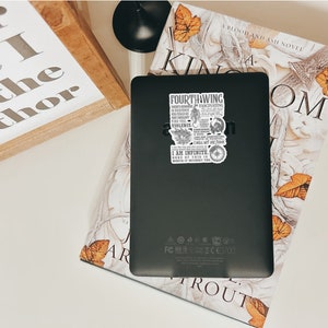 May include: A black e-reader with a sticker on the back. The sticker is a quote from a book called "Fourthing" and features a dragon, an eye, and a sword. The e-reader is sitting on top of a book with a floral design and the title "A Blood and Ash Novel" on the cover.