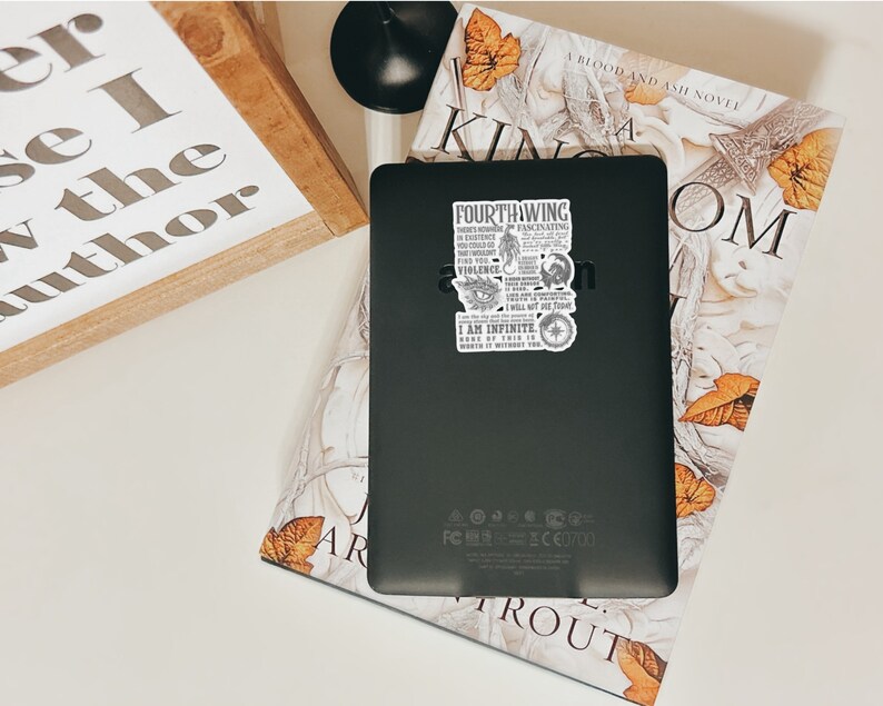 May include: A black e-reader with a sticker on the back. The sticker is a quote from a book called "Fourthing" and features a dragon, an eye, and a sword. The e-reader is sitting on top of a book with a floral design and the title "A Blood and Ash Novel" on the cover.